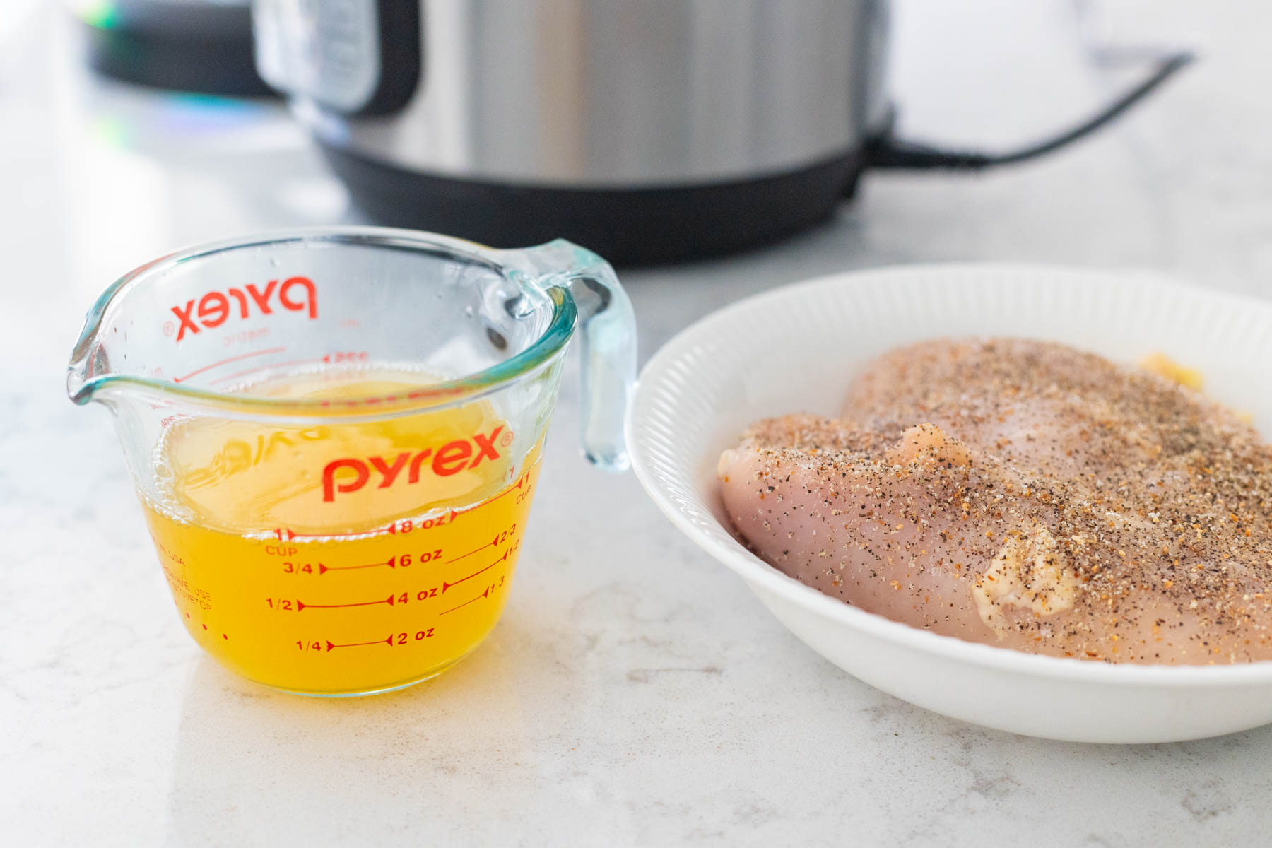 The chicken breasts have seasoning sprinkled over the top and are sitting next to a cup of chicken stock.