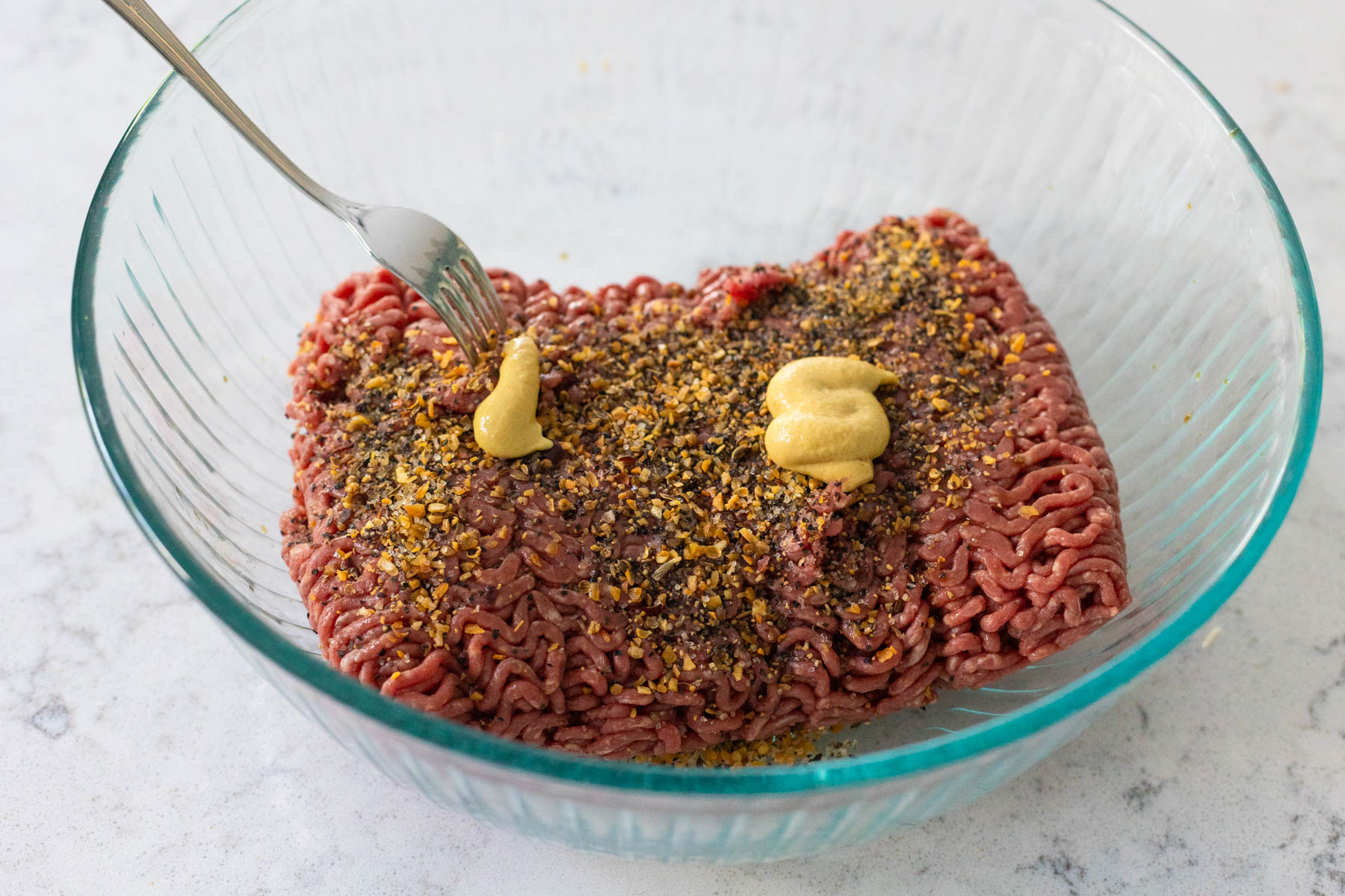 The ground beef is in a mixing bowl topped with seasonings and mustard.