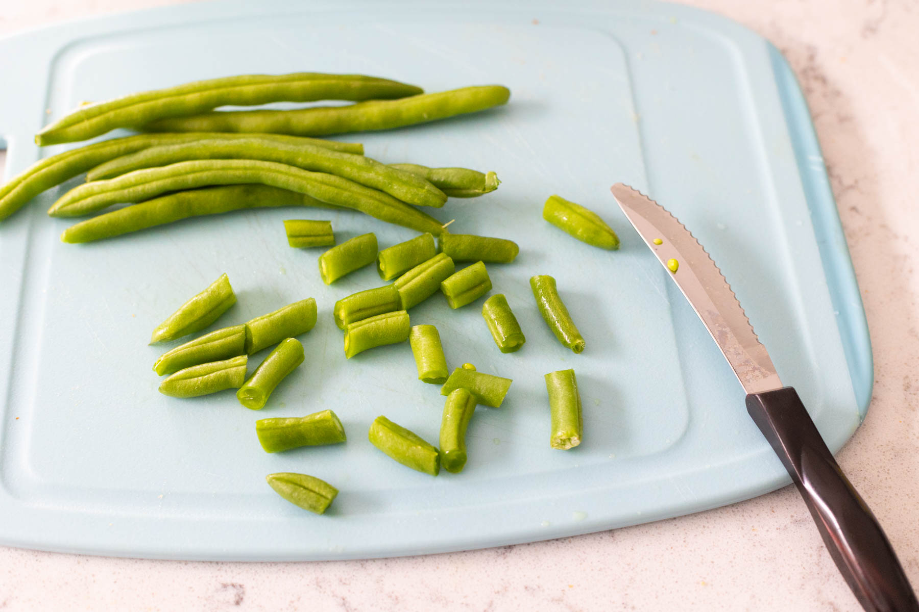The raw green beans are on a cutting board being cut into 1-inch pieces.