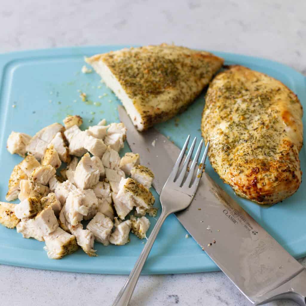 The chicken breasts are on a cutting board, one has been cut into half and chopped into a big dice with a chef knife and fork.