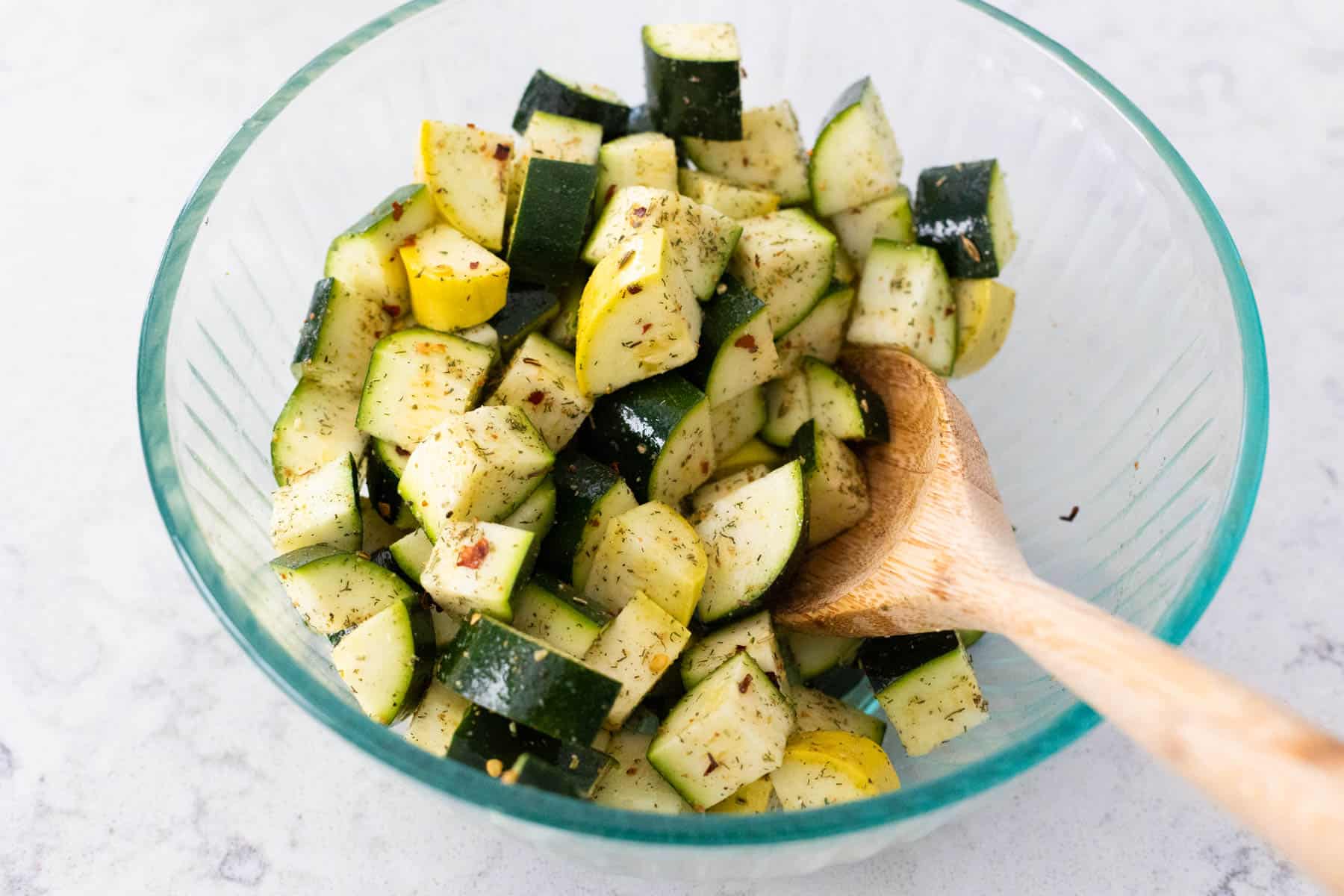 The chopped zucchini and yellow squash are in a mixing bowl coated in olive oil with seasoning on top.