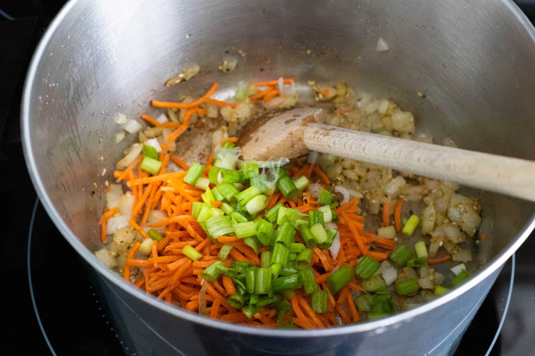 The onions, shredded carrots, minced garlic, and green onions are being cooked in a large soup pot.