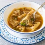 The white bowl is filled with potsticker soup. Half a dumpling is resting on the spoon to show the filling and the peas.